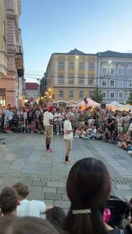 Two young men stand in a public square, facing each other. One, wearing a red cap, white t-shirt, and khaki shorts, juggles three white balls. The other, with blonde hair and wearing a white t-shirt and khaki shorts, watches him. They are surrounded by a large crowd of spectators sitting and standing on the cobblestone ground, watching the performance. Buildings with multiple stories, some with yellow and grey facades, line the square. Tents are visible in the background. The sky is a clear twilight blue. The perspective is from behind a few spectators, looking over their shoulders at the performance.