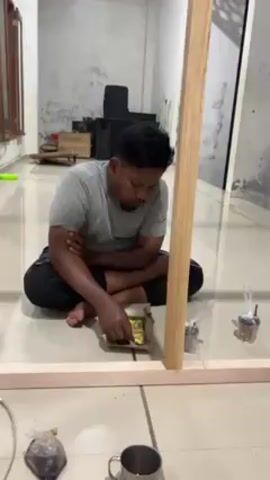 The video shows two men sitting cross-legged on a tiled floor in separate scenes, each inspecting and adjusting the contents of small, wrapped food parcels placed within a rectangular wooden frame. One man wears a gray t-shirt, while the other wears a white t-shirt and a black cap.  Around the frame are metallic cups and other packages. In the background, we can see a window, a doorway, and some wooden boxes, along with an apparent speaker. The second scene, shot at night, also reveals a prayer rug. The focus is on their interaction with the food parcels.