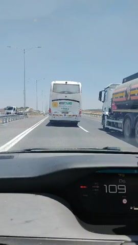 The video shows a view from inside a car driving on a highway. The car's dashboard is visible, displaying the speed as 111 km/h, then 113 km/h, and finally 115 km/h. A white bus with green and yellow lettering is in front of the car. To the right of the bus, a white truck with a tanker trailer is also on the highway. The weather is clear and sunny, and the road appears to be a multi-lane highway with light traffic. The car is in the right lane, and the bus is also in the right lane. The truck is in the lane to the right of the bus. The video captures the car overtaking the bus and moving past it.