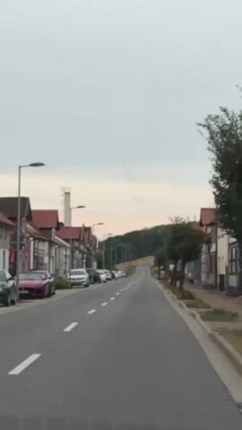A vertical video captures a view from inside a vehicle driving through a village or small town, looking down a street. The street is paved with asphalt and marked with white lane lines. On the left side of the street, there is a row of houses with red tiled roofs and light-colored facades. Several cars are parked along the street. Streetlights are visible along the sidewalk. On the right side of the street, there are also houses and trees. The street curves and ascends a hill, leading towards a densely forested area. The sky is overcast with a pale gray and white hue, suggesting a cloudy or twilight day. The overall scene depicts a quiet, suburban or rural road environment.
