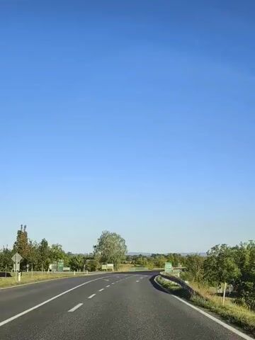 The video shows a point-of-view recording from a vehicle traveling on a road. The road is paved and marked with white lane lines. There are directional signs visible on the side of the road. The surrounding environment consists of green trees and fields. The sky is clear and blue. The time stamps indicate the recording is approximately 5 seconds long.