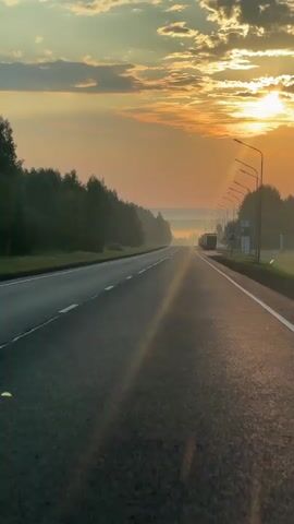 The video shows a view from a moving vehicle on a highway at sunrise. The sun is low in the sky, casting a warm orange glow over the scene. Rays of sunlight are visible, creating a lens flare effect. The road is asphalt with white lane markings. On the left side of the road, there are trees and a foggy mist. On the right side, there are streetlights and more trees. In the distance, several trucks are visible on the road. The sky is filled with clouds that are illuminated by the sunrise. The overall mood is peaceful and serene.
