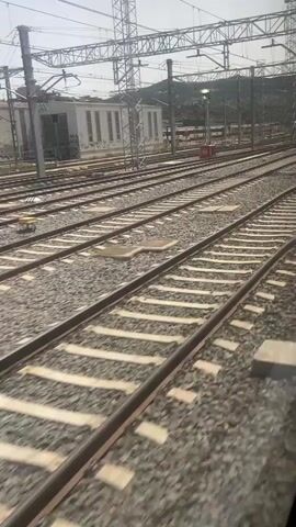 The video shows a view from inside a moving train, looking out at multiple parallel train tracks. The tracks are covered in gravel and sleepers. Overhead power lines and supporting poles are visible. In the distance, there are buildings and hills. The train appears to be in a railyard or station area.