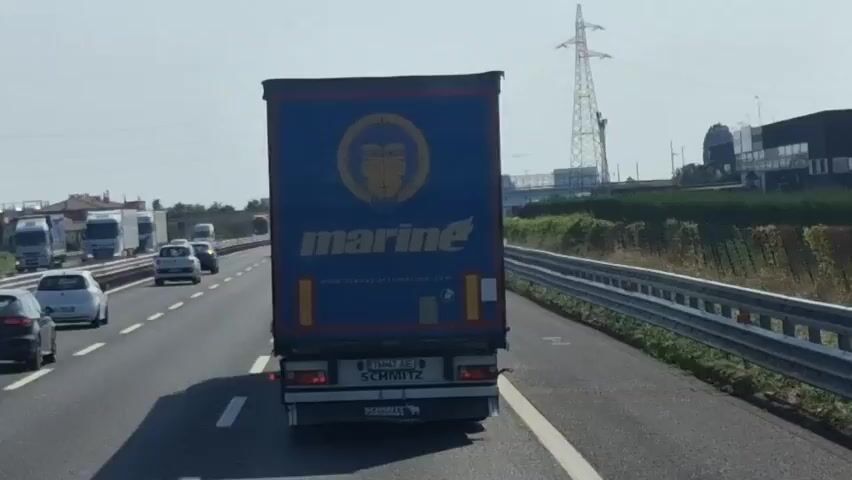 The media file shows a view from inside a vehicle traveling on a highway. The primary subject is the back of a large blue semi-trailer truck with a yellow circular logo and the word "marine" written in white text. The truck also has "SCHMITZ" written on the back. Several other vehicles, including cars and other trucks, are visible on the highway ahead and to the left. The scene is set on a sunny day, with a clear sky and good visibility. The highway has multiple lanes and a metal guardrail on the right side. Beyond the guardrail, there is vegetation and some industrial buildings in the distance. A tall electricity pylon is also visible. The overall impression is of normal daytime highway traffic.