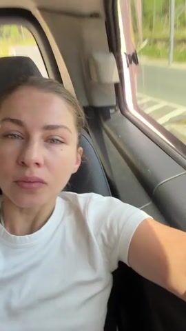This video captures a young woman inside a vehicle, seemingly a car or van, during the daytime. She is wearing a plain white t-shirt. The interior of the vehicle is visible, including dark-colored seats and trim.

The woman is positioned in the foreground, looking directly at the camera with a neutral expression. Her hair is pulled back, and she is wearing subtle makeup.

The background provides glimpses of the outside environment through the vehicle's windows. The scenery suggests a rural or suburban setting with lush green trees and vegetation. The road appears to be winding, indicating movement through a scenic area. The sky is bright and shows patches of white clouds, suggesting fair weather.

The activity appears to be a casual journey, with the woman either filming herself or being filmed during transit. The scene conveys a sense of relaxed travel. There is no visible text in the media. The setting suggests a journey through an area that is somewhat remote, with hints of infrastructure like road signs and possibly a fence in the distance. The overall mood is peaceful and observational.