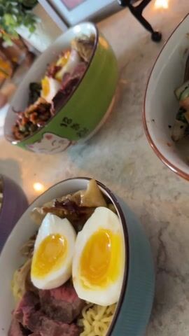 The scene is a close-up shot of three bowls of what appears to be noodle dishes, possibly ramen. Each bowl is a different color, including blue, green, and purple. The blue bowl in the foreground contains noodles, sliced beef, dumplings, and two halved, soft-boiled eggs. The green bowl, positioned slightly behind and to the left of the blue one, contains a mix of ingredients including kimchi, vegetables, beef, and a fried egg. The last bowl, a purple one, is on the left side of the frame and contains beef and noodles. In the background, there is a television screen showing a moving image, a small potted plant, and a framed picture. The bowls are placed on a marble-like surface. Upbeat music is playing in the background.