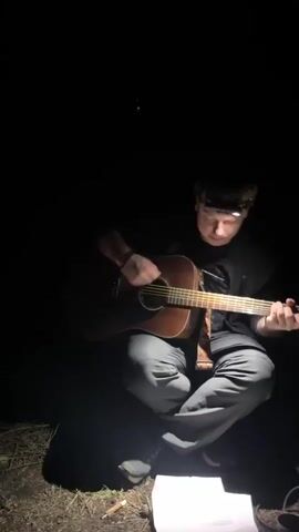 A young man with a headlamp sits cross-legged on the ground in a dark outdoor setting and plays an acoustic guitar. A sheet of paper with text on it is visible near his feet. The headlamp casts a bright light on his face, and he smiles while playing. The surrounding environment appears to be a wooded area at night.