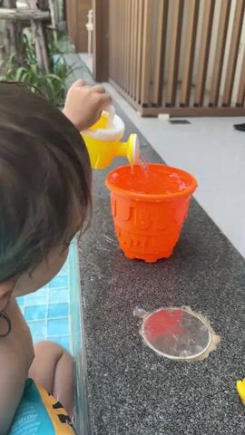 A toddler with short brown hair is seated at the edge of a pool, wearing an orange life vest. The pool water is blue and tiled. The toddler is pouring water from a small yellow and white watering can into a filled, bright orange bucket sitting on the grey stone pool surround. The water overflows as it is poured in. A small yellow rubber duck sits next to the bucket. A stainless steel drain is visible near the bucket. A wooden slat fence and indoor/outdoor furniture are in the background.
