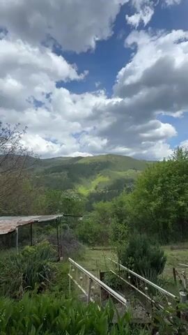 The video presents a static shot of a verdant, mountainous landscape in Rebrovo, Bulgaria, under a partially cloudy sky. In the foreground, lush green plants, including what appear to be lily of the valley, fill the lower left of the frame. To the right, white railings or supports lead up a slight incline. A rustic, weathered shed-like structure with a corrugated metal roof is visible on the mid-left, partially obscured by trees.

Beyond the immediate foreground, a valley unfolds, densely covered in various shades of green trees and foliage, suggesting a rural or semi-wild environment. In the distance, rolling, tree-covered mountains dominate the horizon. Patches of sunlight illuminate parts of the hillsides, creating a dynamic play of light and shadow, indicating the sun is breaking through the clouds. The sky is a mix of bright blue patches and large, fluffy white and grey cumulus clouds. No people or visible text are present in the video. The scene depicts a serene and natural setting, likely captured on a pleasant, mild day.
