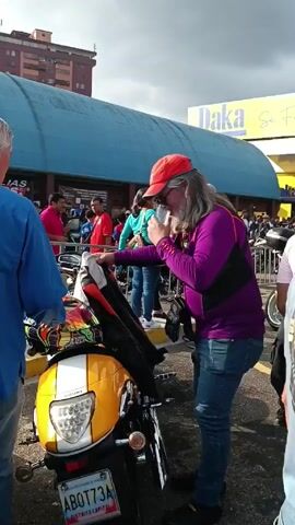 The video shows a man with long grey hair and a beard, wearing sunglasses, a red cap, a purple long-sleeved shirt, and jeans, standing next to a yellow and white Suzuki motorcycle. He holds a clear plastic cup in his right hand and puts his left hand on the motorcycle's seat. A black bag hangs across his chest. 
To his left, another man in a blue shirt stands facing him. Behind them, a crowd of people is visible, along with other motorcycles. In the background, there is a large blue building with the word "Daka" and part of an advertisement on it. A colorful helmet sits on the motorcycle. The license plate is visible, reading "ABOT73A".
The scene appears to be an outdoor gathering of motorcycle enthusiasts.