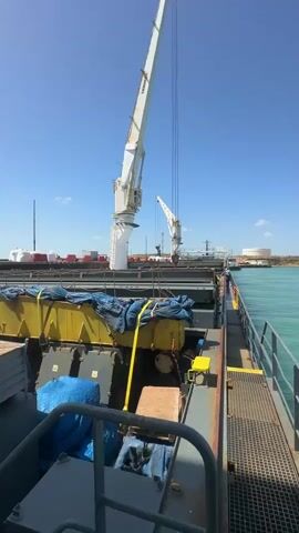 The video presents a wide shot of a ship's deck on a clear, sunny day at a port. The view is from the deck, looking towards the ship's midsection and the port facilities beyond.

A large, white marine crane, with its boom extended, dominates the left side of the frame, hinting at ongoing or recent cargo operations. Another smaller crane is visible further in the background. The sky is bright blue with only a few wispy clouds.

On the ship's deck, several large, yellow cargo units are prominently displayed, covered with blue tarpaulins and secured with yellow straps. One of these units clearly shows the text "SWL 120T", indicating a Safe Working Load of 120 tonnes. Other items, including wooden crates, are also visible on the deck. The ship's structure includes sturdy metal railings and a grated walkway along its side. A lone individual, likely a crew member, can be seen walking on the deck near the railings in the background.

The background reveals a bustling port environment with numerous white containers, trucks, and large storage tanks or silos on the land. The water beside the ship is a distinctive greenish-blue hue. The bright sunlight and clear visibility indicate it is daytime.