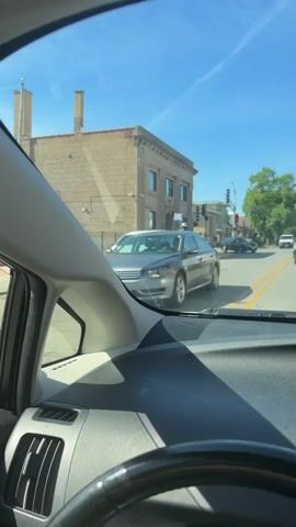 The video shows a man getting into a white minivan on a city street. The man is wearing a blue shirt, dark pants, and a beige bucket hat. He opens the driver's side door of the minivan and leans inside. The minivan is parked on the right side of the street, with a building made of tan bricks visible in the background. A car is parked behind the minivan. Traffic lights are visible at the crosswalk. The video is shot from inside another car. The day is sunny with a clear blue sky.