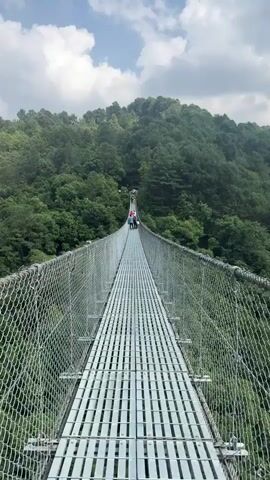 The video shows a long, narrow suspension bridge extending over a lush green valley. The bridge is made of metal grids with wire mesh sides. Several people are visible walking on the bridge in the distance. The surrounding landscape is a dense forest covering the hills, with a few buildings visible in the distance. The sky is partly cloudy. The camera moves, panning from the bridge to the surrounding scenery and then focusing on the metal grids underfoot as a person walks. A foot wearing a white sneaker with pink and black leggings is visible walking on the bridge.