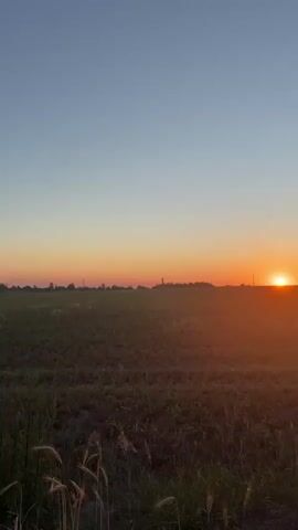 The video shows a sunset over a rural landscape. The sun is a bright orange orb sinking towards the horizon, casting a warm glow across a field of dry grass. Silhouettes of distant trees and a few telephone poles are visible against the colorful sky, which transitions from a pale blue at the top to a deep orange near the horizon. The camera is positioned low to the ground, giving the impression of standing in the field. The image is slightly grainy and there is a lens flare effect from the bright sun.