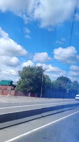 The video shows a bright, sunny day with a blue sky and fluffy white clouds. The footage appears to be taken from inside a vehicle, looking out at a highway. On the highway, several vehicles are visible, including a white van, a white SUV, a white minivan, a white semi-truck with a trailer, and a dark-colored sedan. A sound barrier and trees line the side of the highway. There are power lines visible against the sky. The overall scene is one of normal daytime traffic on a highway.