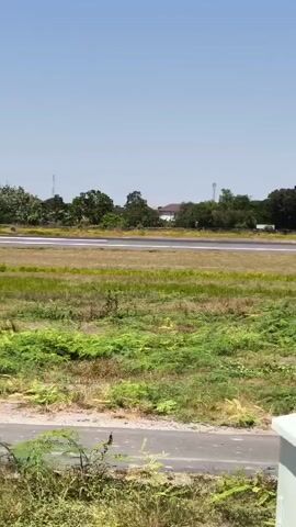 A Garuda Indonesia airplane is taxiing on an airport runway on a bright, clear day. The plane is white with a blue and teal tail fin. The runway is visible in the background, surrounded by green grass and some trees. In the foreground, there are some electrical poles and boxes, as well as some foliage and a white structure. The camera is positioned at a low angle, looking up at the airplane.