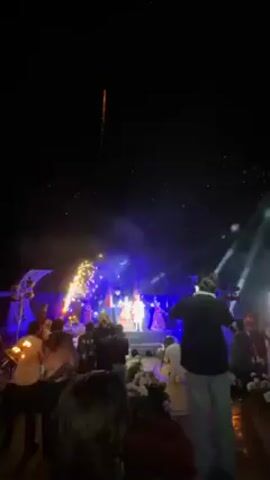 A video captures a celebration at night.  A stage is lit with spotlights. Fireworks explode above it. There are people on the stage, and a crowd of people are standing or kneeling in front of the stage. They are looking at the stage and taking pictures. The scene is punctuated with bursts of fireworks.