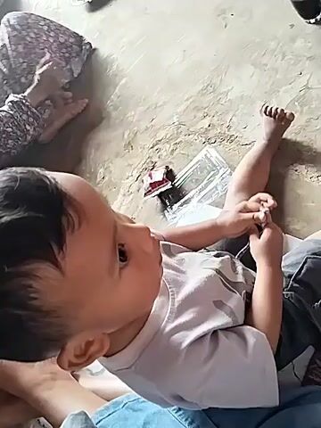 A child is lying on their back on the floor. The child is holding something in their right hand. The child is wearing a white t-shirt and gray pants. There are other people in the background. The environment appears to be indoors.