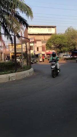 The video shows a truck's perspective of driving past a circular median with a palm tree and a fence. Buildings are visible in the background. The truck is moving forward.
