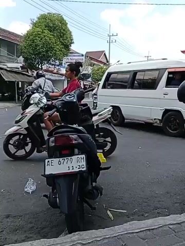 A street scene with several motorcycles and a white van. In the foreground, a black motorcycle is parked facing away from the camera, its license plate reading "AE 6779 RM". To the left, another motorcycle moves away from the camera. In the background, a white van is driving past. Further back, a few other vehicles and buildings are visible, along with trees and power lines against a cloudy sky. A person on a motorcycle wearing a pink headscarf is visible in the middle ground. Another person on a motorcycle wearing a black helmet is visible on the right side of the frame, moving away from the camera. The road appears to be asphalt.