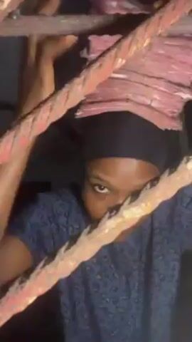 A person holds a large stack of pink money on their head while peering through a metal gate-like structure. The person has dark hair covered by a black band and wears a blue patterned shirt. They are looking directly at the camera. The rusty metal bars of the gate frame the shot. The person's hands are visible, holding the stack of money in place on their head.