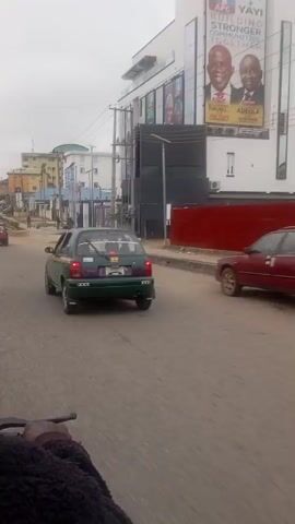 The video is a first-person perspective from a moving vehicle, likely a motorcycle, showing a street scene. The street is paved and appears to be in an urban or semi-urban area. Several vehicles are visible, including a green car in the foreground, a red car further down the road, and other cars in the distance. Pedestrians are also present on the sidewalks and crossing the street. Buildings line the street, with some appearing to be commercial establishments or residential buildings. There are power lines and streetlights visible. The sky is overcast. The camera is moving forward, suggesting the vehicle is traveling down the street. There are no obvious signs of distress or unusual activity, just a normal street scene.