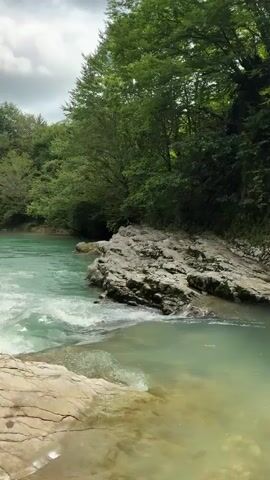 The video shows a flowing river with turquoise water and a rocky, tree-lined shore. The camera pans across the river, showcasing its clear waters and the surrounding lush greenery. There are no people or animals visible in the video. The environment appears to be a natural, serene setting.