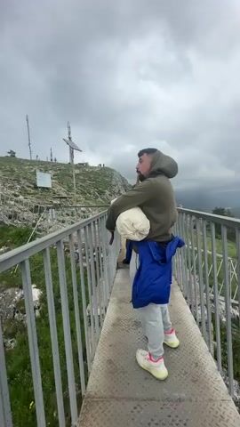 The video shows a man standing on a metal grated bridge, looking off towards a hilltop. The man is wearing an olive green hooded sweatshirt, light gray sweatpants, and yellow and pink sneakers. He has a white object tucked under his arm and a wooden cane in his hand. A blue jacket is tied around his waist. The camera pans to the left to show a series of metal communication towers and a solar panel on the hilltop. The sky is cloudy and overcast. Other people can be seen on the hilltop in the distance. The bridge has a gray metal railing. There is green vegetation and rocks on the hillside below the bridge. The bagpipe music playing in the background is prominent.