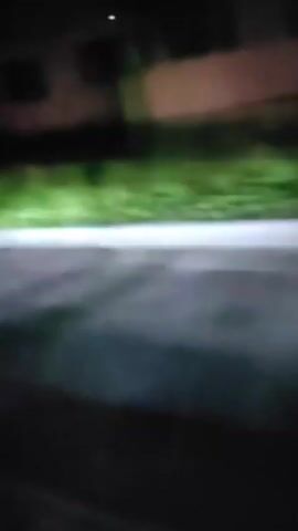 A video taken from inside a car at night shows a blurry view of the road and a grassy area illuminated by the car's headlights. The camera then pans to reveal a smiling woman wearing glasses and a white shirt, seated in a car with a red interior and a seatbelt. She gives a thumbs-up gesture.
