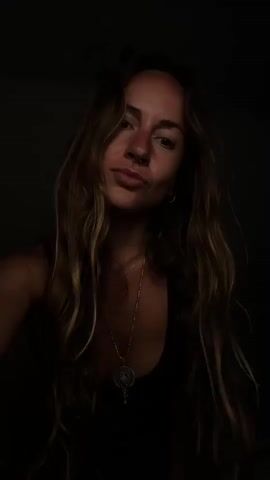 A woman with long, wavy, brown hair with lighter highlights is featured in a close-up, chest-up shot. She wears a dark top, possibly a tank top, and a silver-colored necklace with a round, ornate pendant and a smaller charm. A septum piercing is visible.

The lighting is low-key, illuminating her face and upper body from the front, while the background remains completely dark, creating a dramatic effect. She looks directly at the camera with a calm, slightly smirking expression.

Throughout the short video, she interacts subtly with the camera. Initially, she holds a slight smile, then moves her hands to pull her long hair forward over her shoulders, framing her face. Her hands then briefly rest near her chest/shoulders before the clip ends. There is no visible text. The setting is indoors, and the context provided indicates the location is Graz, Austria.