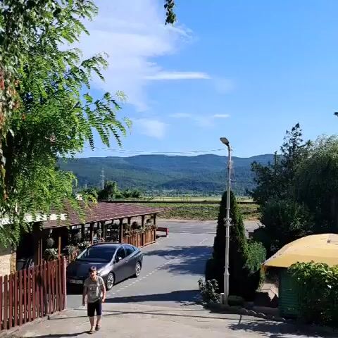 A boy runs down a paved pathway in front of a building with an outdoor seating area and a fence. A dark sedan is parked to the right of the building. Further in the background, a dark blue van is parked next to a large evergreen tree. The scene is set against a backdrop of rolling hills and a bright blue sky with scattered clouds. The pathway is lined with trees and a stone wall.