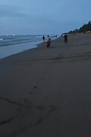 The video shows a beach scene at dusk. The tide is out, leaving wet sand near the water's edge. Several people are present, some walking, and at least two are riding motorcycles on the beach. There are some distant lights visible on the water, possibly boats. The sky is cloudy and a deep blue, indicating the transition from day to night. The overall atmosphere is serene and tranquil.