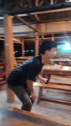 A young man is featured in an outdoor or semi-outdoor setting, possibly a restaurant or cafe, at night. He is wearing a black t-shirt with "WOOPIE PROJECT" printed on it, and dark jeans. The setting includes wooden tables and benches, with artificial lighting illuminating the area and distant lights visible in the background.

The video begins with the man in a slightly crouched position, then he stands upright. He holds a small black tube or bottle, which appears to be a product. He turns towards the camera, points at the bottle, and seems to be talking, possibly presenting or demonstrating the product. The visible text on the bottle includes "morris" and "OIL CONTROL +". On one of the wooden tables, the number "12" is visible. The man has a engaged expression, indicating he is actively communicating.