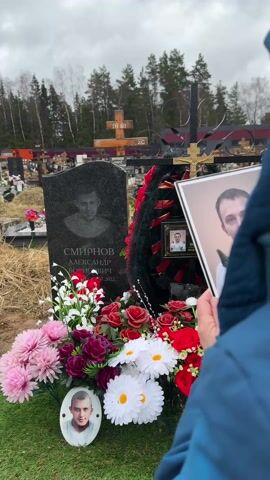 A person is standing at a gravesite in a cemetery. The grave has a dark grey headstone with a carved image of a person. In front of the headstone, there are colorful artificial flowers, including pink chrysanthemums, red roses, and white daisies. On the ground is a framed photo of a young man. Behind the headstone is a black iron cross with a gold crucifix, adorned with a wreath featuring black, red, and orange ribbons, and another framed photo. The person, wearing a blue jacket, is holding a framed photo of a man. The cemetery has numerous other gravesites visible in the background, along with trees. The sky is overcast.