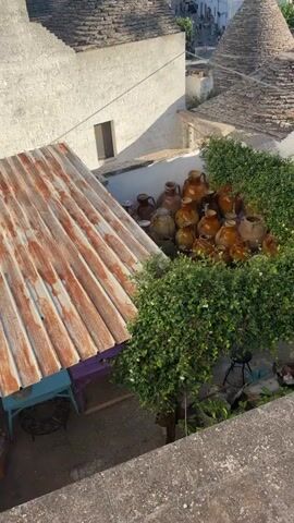 The video shows a high-angle view of a village with conical-roofed buildings, likely in Alberobello, Italy. In the foreground, there is a rusty corrugated metal roof. To the right of this roof, there is a collection of terracotta pots and jugs, partially obscured by green foliage. Beyond the pots, a conical stone roof is visible. Further in the background, the characteristic white, conical-roofed houses of the village stretch across the landscape under a clear blue sky. There are a few people visible walking in the distance. The overall scene depicts a charming, sunlit view of the unique architecture of the region.