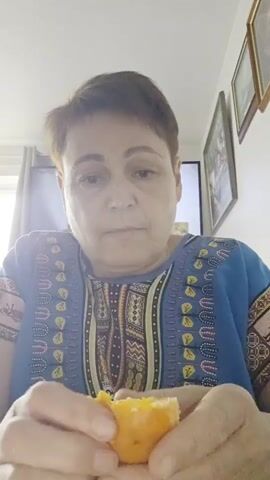 A close-up video shows a middle-aged woman with short brown hair, wearing a blue top with an ethnic pattern, peeling an orange with both hands. She looks directly at the camera with a neutral to slightly pleasant expression, breaking into a small smile around the 0:04 mark.

The setting appears to be indoors, likely a living room or dining area in a home. In the background, natural light enters from a window on the left. A television screen is visible behind her, along with several framed pictures on the wall to the right. Text is visible on the television screen, which appears to be in a foreign language (possibly Polish), with phrases like "takich interierych" and "takich" discernible. The overall lighting suggests it is daytime. The activity is simple and domestic, focusing on the woman's interaction with the fruit.