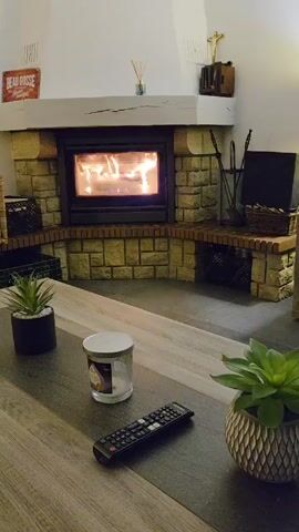 The camera slowly pans across a living room scene, showing a stone fireplace with a roaring fire, a coffee table with two small potted plants, a candle in a glass jar, and a TV remote. The fireplace tools are next to the fire and firewood in a basket to the side. A red sign reads "BEAU GOSSE" above the fireplace.