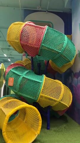 A 4-second video shows a colorful indoor playground. The playground features a large, multi-colored tube slide constructed from yellow, pink, and green netting. The slide is supported by blue poles and winds its way through the space. Adjacent to the slide is a tall, green net climbing structure with multiple levels. The walls of the playground are decorated with vibrant murals depicting fantastical plants in shades of green, blue, and purple. The floor is covered in green artificial turf. In the background, a large, silver tube slide descends towards a pile of colorful building blocks. The ceiling has overhead lighting. The video pans slightly to the right, revealing more of the playground's features.