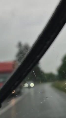This is a vertical video of a car driving on a wet, paved road on an overcast day. The road curves to the right, with the surrounding landscape consisting of lush green trees and vegetation. The sky is a muted gray, suggesting rain or heavy clouds. 

In the foreground, a dark-colored sedan is visible driving in the right lane. Its headlights are on, illuminating the wet road ahead. The road has white double lines marking the lanes. The car appears to be the primary object of focus in the initial frames. Further down the road, another car is visible, also driving in the same direction.

The scene is set in a rural or semi-rural area, indicated by the dense greenery and lack of urban structures. The presence of water droplets on the camera lens adds to the impression of a rainy or misty atmosphere. The overall mood conveyed by the footage is serene and a bit melancholic due to the overcast weather and the solitary nature of the drive.

There is no visible text within the video itself, though a white circular sign is visible on the left side of the road near a large pine tree. Another sign, possibly a billboard, is barely visible in the distance on the right side of the road. The location context provided is Balgarski Izvor, Bulgaria.