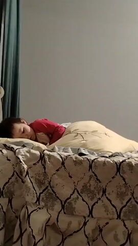 The video shows a child lying in bed. The child is wearing a red top and is covered with a light-colored blanket. The bed has a patterned bedspread. There is a turquoise curtain in the background. Someone off-camera says "Привет," which means "Hello" in Russian, and the child responds, "Привет."