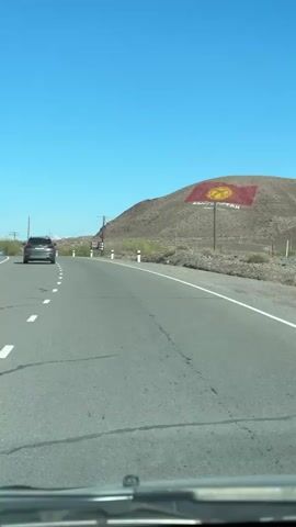 The video shows a road and landscape. The video is taken from the interior of a vehicle traveling on a road.  The sky is bright blue. A car is driving ahead of the vehicle taking the video. On a nearby, treeless hill is a large depiction of the flag of Kyrgyzstan and the word "Kyrgyzstan" is written under it. There are power lines running overhead. A sign is on the side of the road.