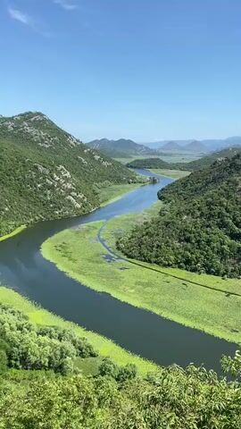 A wide shot shows a river with light green foliage on its banks. The river curves around a large, green mountain in the center of the frame, disappearing behind it. The mountains in the distance appear to be low and covered with green trees. The sky is bright blue and cloudless.