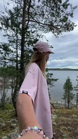 A woman wearing a pink baseball cap and a light pink oversized t-shirt stands on a mossy, rocky terrain overlooking a body of water. She is seen from a first-person perspective, with her arm extended forward. In the background, there are evergreen trees and a cloudy sky. The woman appears to be looking out at the water.