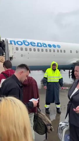 A group of people are boarding an airplane via a mobile staircase. The airplane is white with the word "ПОБЕДА" (Pobeda) written in blue letters and blue dots. A person wearing a high-visibility yellow and blue rain suit stands near the bottom of the stairs, appearing to direct passengers. The ground appears wet, suggesting it may be raining or has recently rained. The passengers are dressed in casual clothing and some are carrying bags. The camera is positioned behind the passengers, looking up the stairs towards the airplane entrance. The sky is overcast and gray.