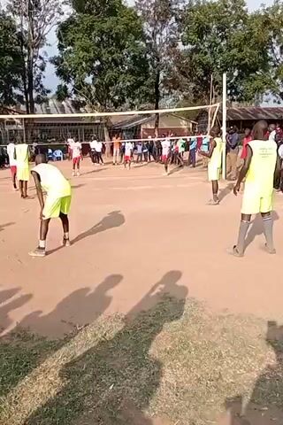 A group of young people are playing volleyball on a dirt court. The court has a net stretched across the middle. Some of the players are wearing neon yellow uniforms, while others are wearing red and white. In the background, there are trees, buildings, and a crowd of spectators. The overall atmosphere appears to be one of casual recreation.