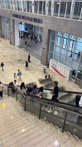 The video shows an interior view of a large, modern building, likely a transportation hub such as a train station or airport terminal. The perspective is from an elevated position, looking down onto a wide, open concourse area. In the foreground, a set of escalators and stairs are visible, with several people ascending. These individuals are casually dressed. Some are alone, while others are in small groups. On the escalators, a few individuals are engaged in conversation, while others appear to be focused on their phones or simply observing their surroundings.

The concourse floor is made of polished concrete or tile. Scattered across this area are a few scattered figures. Some people are walking, while others are sitting or standing in small groups, some with luggage. There are large windows that allow natural light to enter the building. In the background, there is signage in a language that appears to be Chinese. A prominent architectural feature in the background is a large, arched entryway or passage, beyond which more people are visible, suggesting different sections of the terminal. Several white, modular units, possibly lockers or information kiosks, are lined up against a wall to the right. The overall impression is of a busy but orderly public space. The lighting is bright and artificial, typical of indoor public venues. The architecture appears modern and functional, with steel and glass elements prominent.