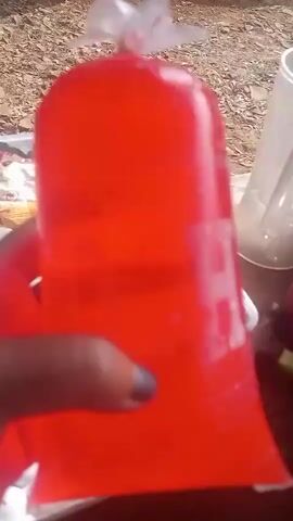 A hand with blue nail polish holds a red, translucent plastic bag filled with red liquid. The bag appears to be sealed at the top with a knot in the plastic. Water droplets are visible on the outside of the bag. In the background, there is a white plastic container with what appears to be ice inside, and other objects that are out of focus. The lighting suggests an outdoor environment.