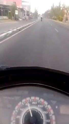 The video shows a first-person perspective of someone riding a motorcycle down a paved road lined with buildings on the left and trees on the right. Several other motorcycles are visible on the road. The sky is bright, suggesting daytime.
