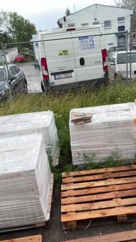The video shows an outdoor scene with stacks of palletized goods wrapped in plastic in the foreground. Wooden pallets are scattered on the ground, some of which are wet and placed in muddy puddles. The ground is a mixture of dirt, gravel, and scattered debris, including bricks and cigarette butts. In the background, a dark-colored car is parked near a construction fence. Behind the fence, a white van is visible, along with a building. The overall environment appears to be a construction site or storage area.