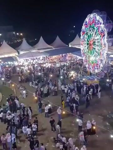 The video shows an aerial view of a nighttime fair or carnival. In the foreground, a large crowd of people, many wearing light-colored clothing and headscarves, are gathered. Several rows of tents with peaked roofs are set up in the midground, illuminated by strings of lights and internal lighting, suggesting vendor stalls or food booths. A prominent, brightly lit Ferris wheel with a colorful, geometric design is visible on the right side of the frame. In the mid-left, a carousel or swing ride with people on it is in motion, also adorned with colorful lights. The ground appears to be a dirt or grassy area, with some scattered debris. The overall atmosphere is festive and crowded, with artificial lighting creating a vibrant scene against the dark night sky.